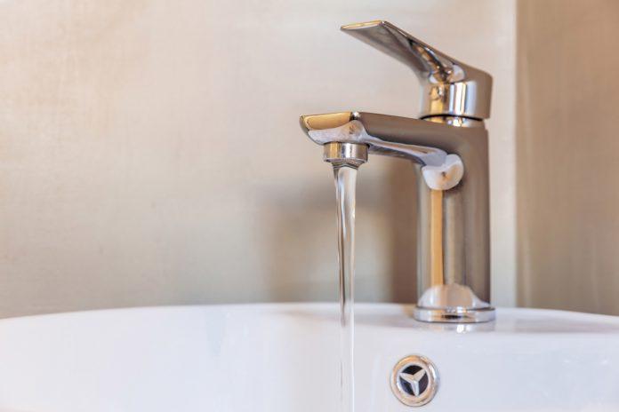 Sink basin faucet, bathroom interior detail. Open chrome tap with running water on white washbasin.